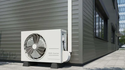 Das Bild zeigt eine außen am Gebäude installierte weiße Wärmepumpen- oder Klimaanlagen-Außeneinheit (Dual Inverter) auf zwei Sockeln. Die Zuleitung verläuft in einem weißen, senkrechten Kabelkanal an der Fassade und führt geschützt in das Gerät.