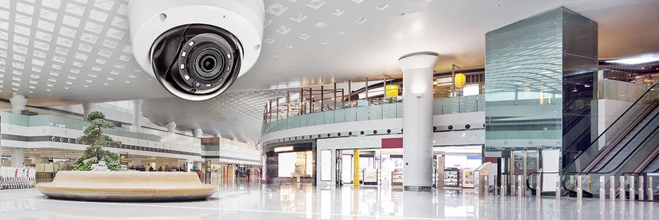 Das Bild zeigt eine Überwachungskamera, die in einer modernen, weitläufigen Empfangshalle oder Lobby installiert ist.