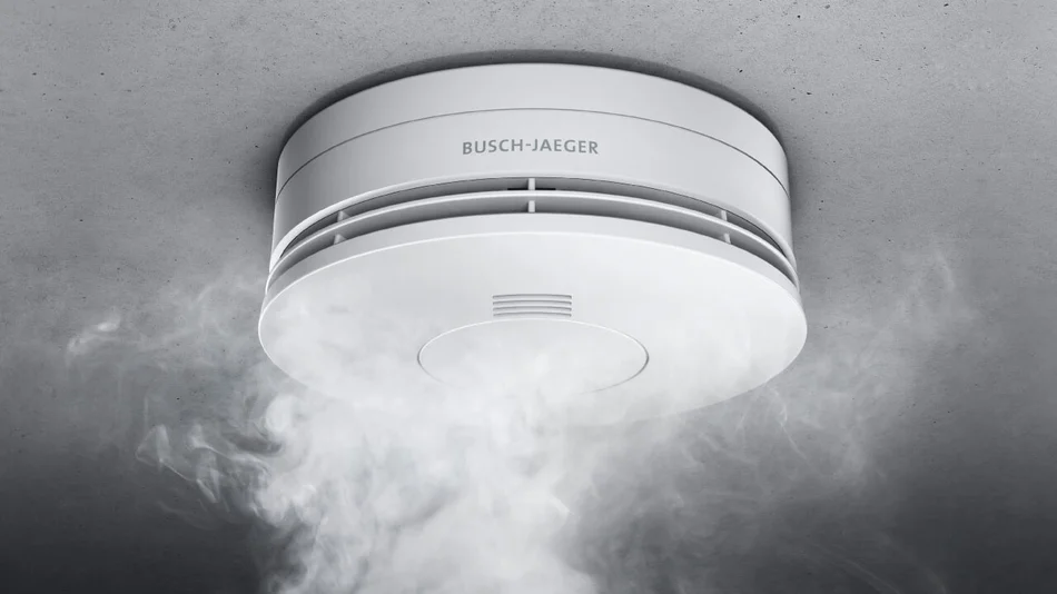 Das Bild zeigt einen weißen Busch-Rauchalarm® Alarm-Detektor an einer grauen Decke. Von unten steigt Rauch auf.