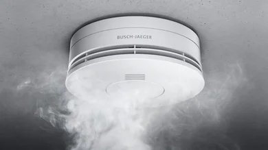 Das Bild zeigt einen weißen Busch-Rauchalarm® Alarm-Detektor an einer grauen Decke. Von unten steigt Rauch auf.