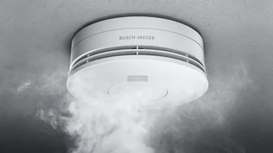 Das Bild zeigt einen weißen Busch-Rauchalarm® Alarm-Detektor an einer grauen Decke. Von unten steigt Rauch auf.