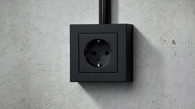 Das Bild zeigt eine Steckdose des Herstellers Jung in der Farbe Schwarz, die an einer Wand montiert ist.