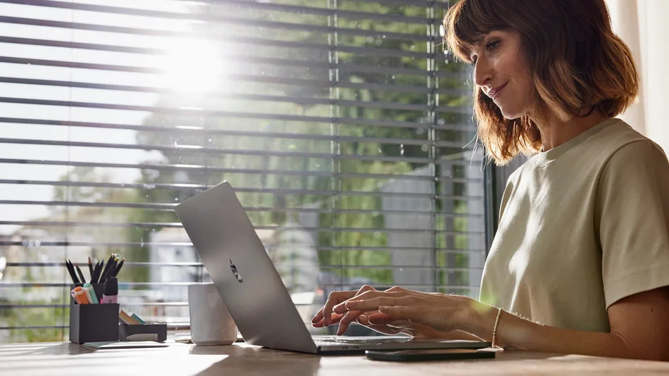 Das Bild zeigt eine Frau, die vor einem Fenster an einem Schreibtisch sitzt und an einem Laptop arbeitet. Am Fenster sind Jalousien heruntergelassen. 