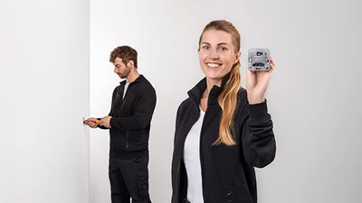 Zwei Personen. Im Vordergrund eine Frau, die einen Dimmer in der Hand hält. Im Hintergrund ein Mann, der gerade etwas an der Wand montiert.