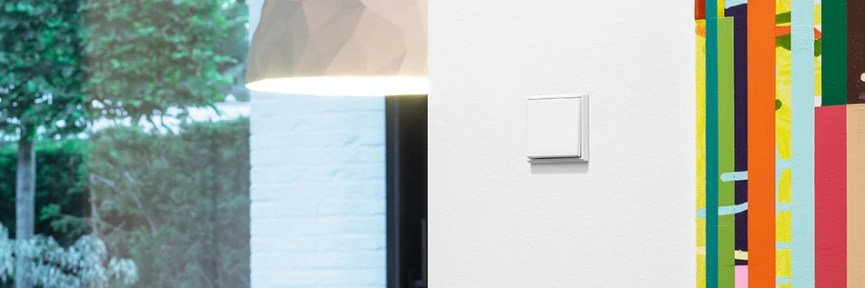 Schalter des Herstellers Jung in der Farbe Weiß an der Wand im Vordergrund. Dahinter Blick auf eine Hängelampe in der Farbe Weiß.