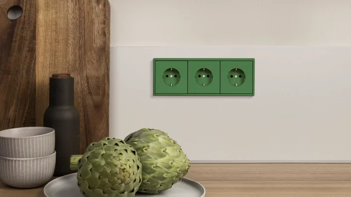Das Bild zeigt eine Dreifachsteckdose in Grün, die in eine weiße Küchenrückwand integriert ist. Auf der Arbeitsplatte davor stehen zwei Artischocken, eine dunkle Flasche und zwei Schalen.