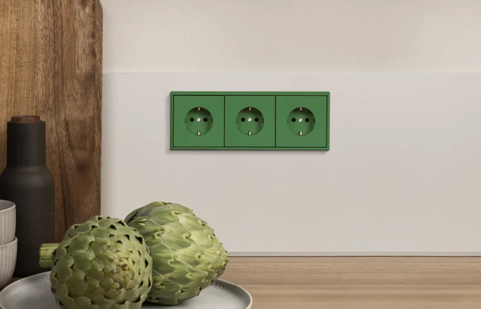 Das Bild zeigt eine Dreifachsteckdose in Grün, die in eine weiße Küchenrückwand integriert ist. Auf der Arbeitsplatte davor stehen zwei Artischocken, eine dunkle Flasche und zwei Schalen.
