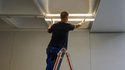 Das Bild zeigt eine Person auf einer Leiter, die eine längliche LED-Leuchte unter einer Raumdecke befestigt. 