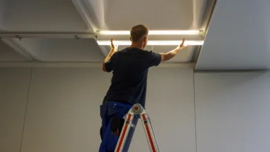 Das Bild zeigt eine Person auf einer Leiter, die eine längliche LED-Leuchte unter einer Raumdecke befestigt. 