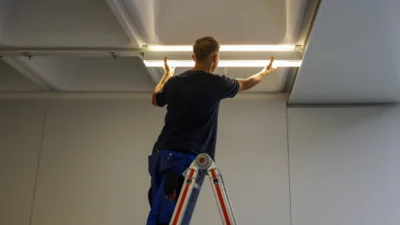 Das Bild zeigt eine Person auf einer Leiter, die eine längliche LED-Leuchte unter einer Raumdecke befestigt. 