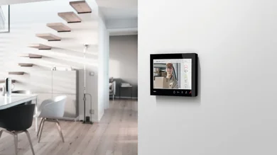 Das Bild zeigt im rechten Bildbereich eine schwarze Wohnungsstation an einer weißen Wand. Im rechten Bildbereich wird ein moderner Wohnbereich mit einem Essbereich gezeigt Eine freischwebende Treppe mit Holzstufen führt ins Obergeschoss.
