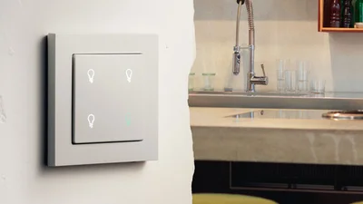 An der Wand montierter großer grauer Tastsensor. Dahinter Blick auf die Spüle einer Küche.