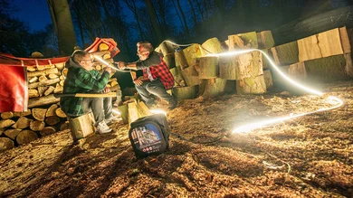 Das Bild zeigt zwei Männer in Holzfällerhemden bei Nacht in einem Wald. Sie sitzen zwischen aufgestapeltem Brennholz und Holzspänen. Eine leuchtende LED-Lichtleiste, die von einer Kabeltrommel am Boden kommt, beleuchtet die Szene und wird von den Männern gehalten.