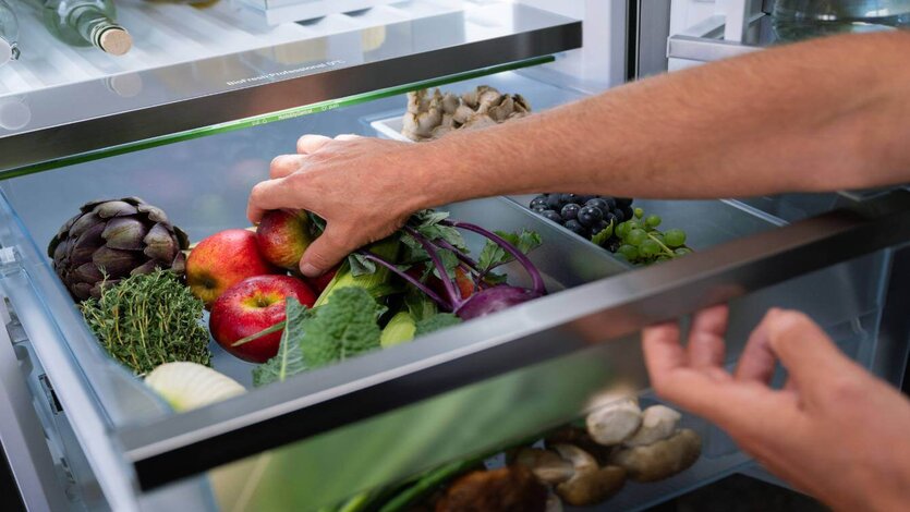 Eine Hand greift in die Gemüseschublade eines Kühlschranks mit frischem Obst und Gemüse wie Äpfeln, Trauben und Artischocken.