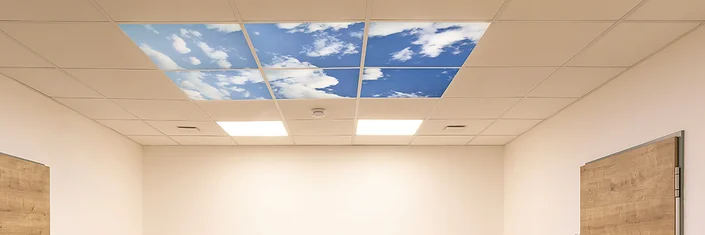 Das Bild zeigt eine Decke mit Deckenpanelen. Sechs Panele zeigen Wolken an einem blauen Himmel. Zwei Panele enthalten Panelleuchten.