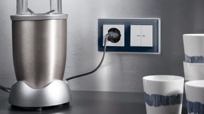 Ein silberner Mixer steht auf einer Arbeitsplatte vor einer gebürsteten Metallwand. Dort ist eine Steckdose mit eingestecktem Kabel und ein doppelter Lichtschalter in einem dunkelblauen Rahmen angebracht. Rechts daneben stehen mehrere weiße Tassen mit blauen Streifen.