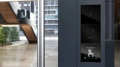 Ein Gira System 106 IP ist an einer dunklen Säule im Eingangsbereich montiert. Im Hintergrund ist eine moderne Architektur mit Glas, Metall und Holz zu sehen.