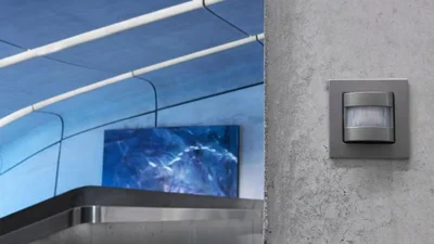 Ein Gira Bewegungsmelder ist an einer grauen Wand montiert; im Hintergrund sieht man einen modern gestalteten Innenraum mit blauen und weißen Flächen.