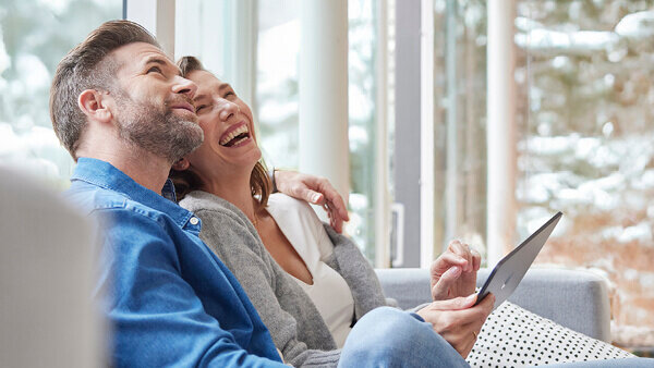 Das ild zeigt ein entspanntes Paar, das gemeinsam auf dem Sofa sitzt und ein Tablet benutzt, mit Blick auf ein großes Fenster und winterliche Landschaft draußen.