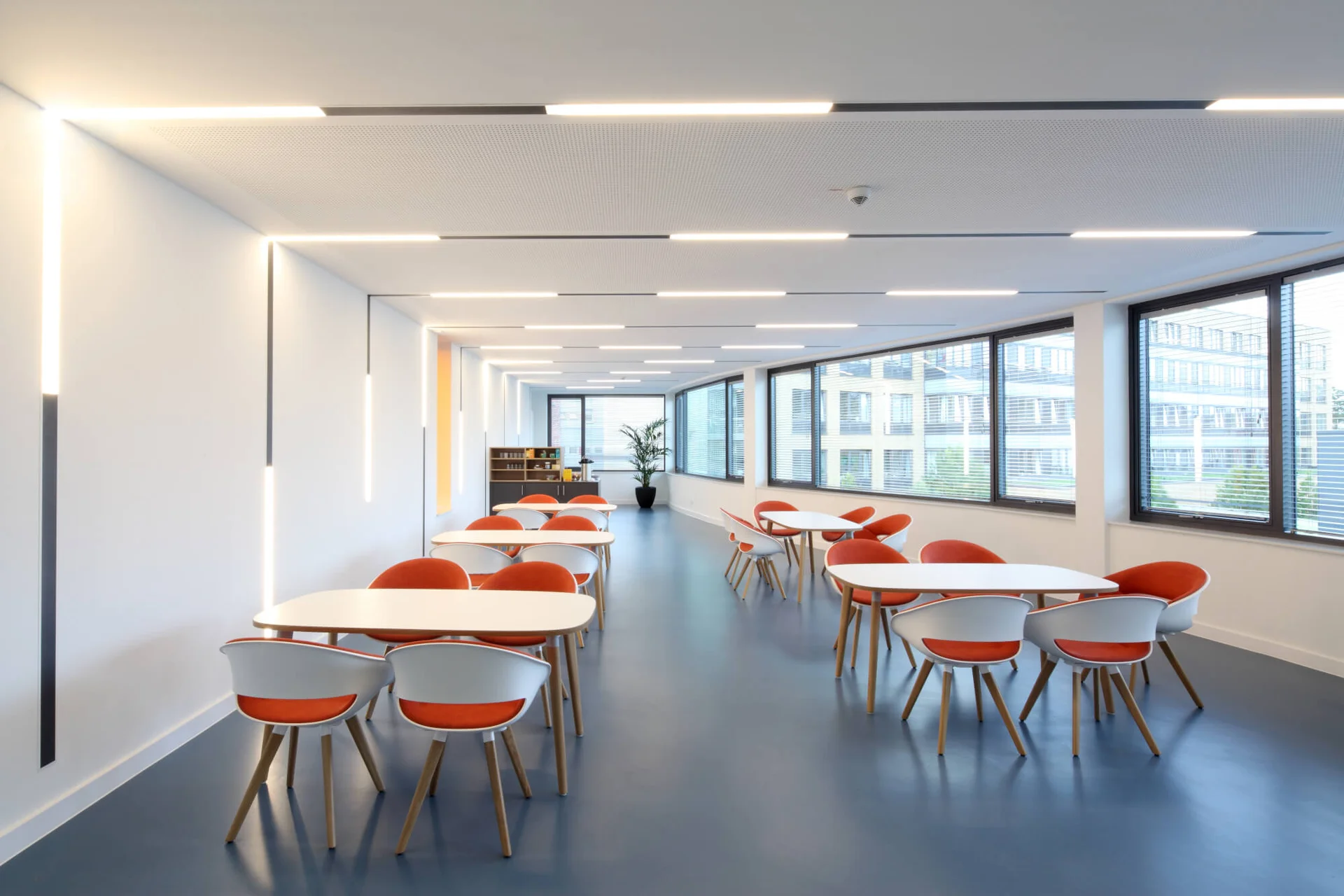 Das Bild zeigt einen hellen Raum mit weißen Tischen und orange gepolsterten Stühlen. Der Boden ist blau, die Decke weiß mit integrierten Leuchten. Links sind vertikale Lichtleisten an der Wand, rechts große Fenster mit Jalousien. Hinten stehen ein Regal und eine Pflanze.