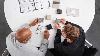 Das Bild zeigt zwei Männer an einem Tisch, die verschiedene Lichtschalter-Designs begutachten. Auf dem Tisch liegen Muster, ein Smartphone mit Produktinformation und ein Bauplan.