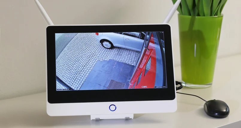 Das Bild zeigt einen Monitor mit integrierter Überwachungstechnik, auf dem das Livebild einer Sicherheitskamera zu sehen ist. Der Monitor steht auf einem hellen Sideboard, daneben befindet sich eine schwarze Computermaus und ein grüner Blumentopf mit Tulpen. Die Kamera überwacht einen Eingangsbereich mit gepflastertem Boden und einem geparkten Auto.