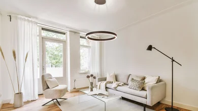 Heller Wohnraum mit Sofa, Sessel, Stehleuchte und Glastisch. Über dem Sitzbereich hängt eine ringförmige Pendelleuchte an feinen Seilen von der Decke.