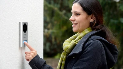 Das Bild zeigt eine Frau, die draußen eine elegante Türsprechanlage mit Kamera und Klingelknopf betätigt.