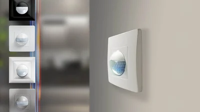 Das Bild zeigt mehrere Wandbewegungsmelder in unterschiedlichen Farben und Designs. Einer der Sensoren ist prominent an einer hellen Wand montiert, während links Varianten in Schwarz, Weiß und Grau gezeigt werden.