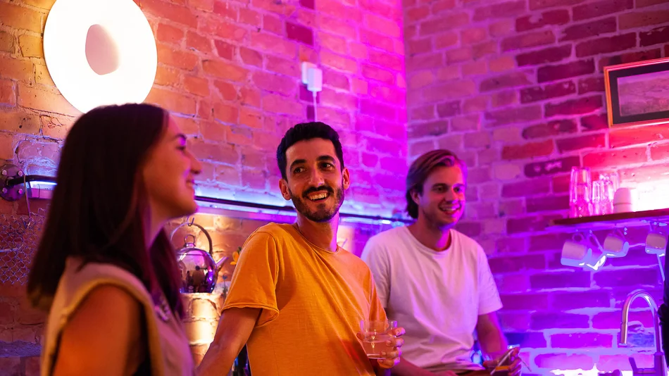 3 Personen in einer Bar, die lachen. Im Hintergrund typische Barbeleuchtung in der Farbe Lila.