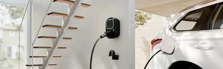 Das Bild zeigt eine Wallbox-Ladestation an einer Wand, die ein weißes Elektroauto lädt, während im Hintergrund eine Treppe und ein Fenster zu sehen sind.