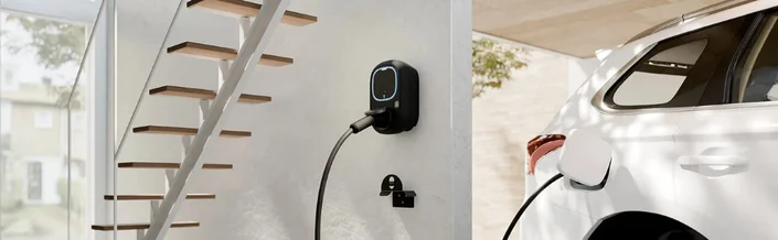 Das Bild zeigt eine Wallbox-Ladestation an einer Wand, die ein weißes Elektroauto lädt, während im Hintergrund eine Treppe und ein Fenster zu sehen sind.