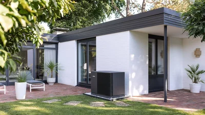 Das Bild zeigt einen modernen, weißen Flachdach-Bungalow mit Garten und Terrasse. Vor einer Hauswand steht eine schwarze Wärmepumpe.