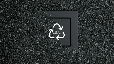 Zu sehen ist eine schwarze Schalterabdeckung, umgeben von vielen schwarzen Kunststoffgranulaten. In der Mitte steht ein Recycling-Symbol mit dem Hinweis „95% Rezyklat“