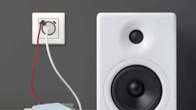 Eine weiße Steckdose mit USB-C-Anschluss ist an einer grauen Wand montiert, wobei ein rotes und ein weißes Kabel zum Laden von Geräten eingesteckt sind. Rechts daneben ist ein Teil eines weißen Lautsprechers mit schwarzem Hoch- und Tieftöner zu sehen.