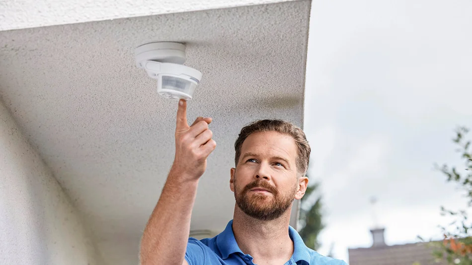 Das Bild zeigt eine Person, die einen Bewegungsmelder mit ihrem Zeigefinger berührt. Der Bewegungsmelder ist an einem Deckenvorsprung im Garten befestigt.
