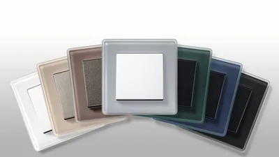 Eine Reihe von Lichtschaltern in verschiedenen Farben, von Weiß über Beige und Braun bis hin zu Grün, Blau und Schwarz, sind fächerförmig auf einem hellgrauen Hintergrund angeordnet. Jeder Schalter hat einen quadratischen Rahmen und einen passenden Schaltereinsatz.