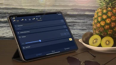 Das Bild zeigt ein Tablet aufgestellt auf einem braunen Tisch. Auf dem Tisch liegt eine Sonnenbrille. Eine Obstschale mit Kiwis und einer Ananas steht neben dem Tablet. Der Hintergrund zeigt Wellen und einen Sonnenuntergang