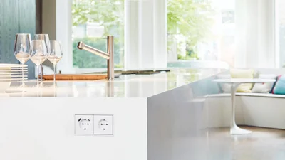Eine moderne Küche mit einer weißen Arbeitsplatte, auf der Weingläser und ein Stapel Teller stehen. Im Vordergrund ist eine weiße Doppelsteckdose mit USB-Anschlüssen zu sehen. Im Hintergrund, durch große Fenster, scheint helles Tageslicht herein, und ein gemütlicher Sitzbereich mit Kissen ist erkennbar.