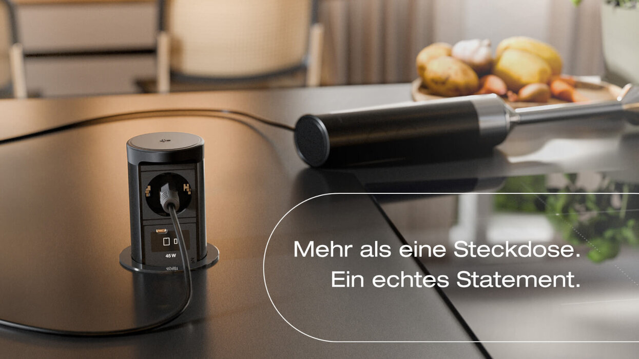Auf einem Tisch steht eine versenkbare schwarze Steckdoseneinheit mit USB-Anschluss und eingestecktem Kabel. Im Hintergrund liegen eine Zange und Obst auf einer Schale, dazu steht der Text „Mehr als eine Steckdose. Ein echtes Statement.“