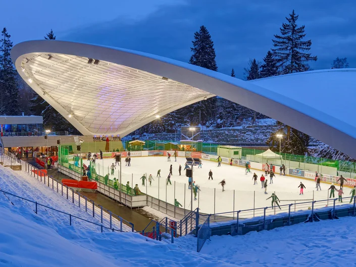 Das Bild zeigt eine überdachte Eisbahn im Freien, auf der viele Menschen Schlittschuh laufen. Um die Eisfläche herum befinden sich Zäune, Gebäude und Bäume im Hintergrund.