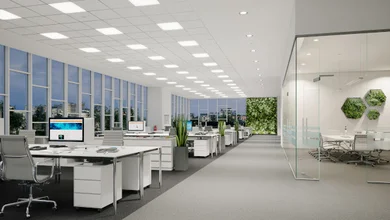 Großraumbüro mit vielen Schreibtischen, großen Fenstern und Deckenleuchten.