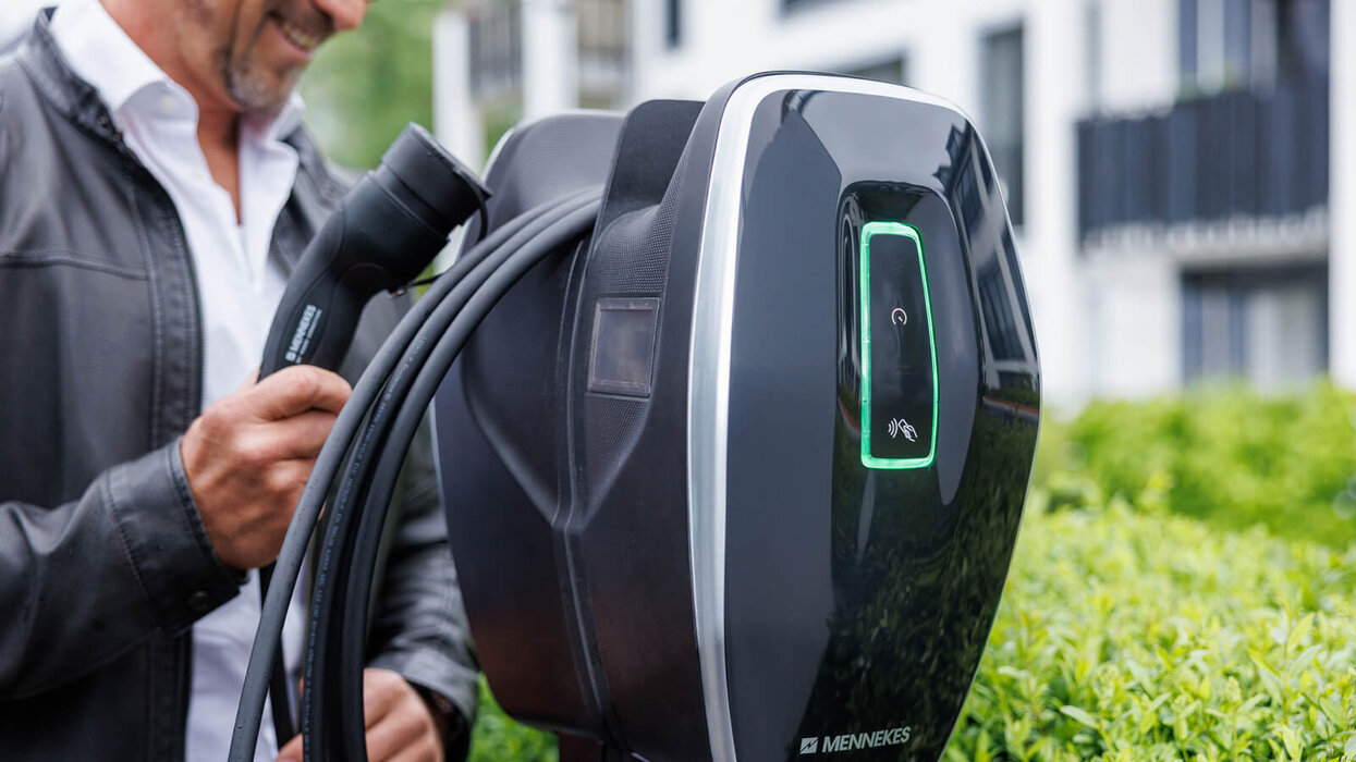 AMTRON® 4Business 700 von Mennekes Ein Mann steht neben einer schwarzen Ladestation für Elektroautos und hält ein Ladekabel mit Stecker in der Hand. Die Station zeigt ein grün leuchtendes Symbol. Im Hintergrund sind moderne Gebäude und grüne Pflanzen zu sehen.