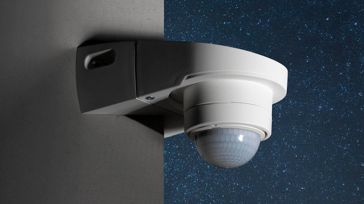 Das Bild zeigt einen weißen Bewegungsmelder, der an einer Wand montiert ist. Der Hintergrund ist zur Hälfte dunkelblau mit Sternen, zur anderen Hälfte grau.