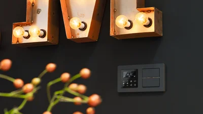 Das Bild zeigt eine dunkle Wand, an der ein modernes Smart-Home-Bedienfeld mit digitaler Anzeige und Tasten montiert ist. Darüber befinden sich beleuchtete Vintage-Buchstaben aus Metall, die mit Glühbirnen versehen sind. Im Vordergrund ist unscharf ein Zweig mit rötlichen Beeren oder Knospen zu sehen, der dem Bild eine natürliche Note verleiht.