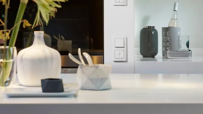Zu sehen ist eine elegante Küchen- oder Wohnsituation mit einem weißen JUNG-Schalter an der Wand. Im Vordergrund stehen dekorative Gefäße und auf der rechten Seite Flaschen und Glasbehälter auf einer Ablage.