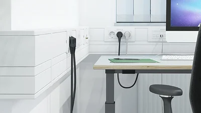 Helles Büro mit weißem Schreibtisch, Monitor auf dem Tisch und schwarzem Bürostuhl nebst Geräteeinbaukanal von OBO Bettermann links an der Wand.
