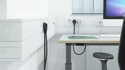 Helles Büro mit weißem Schreibtisch, Monitor auf dem Tisch und schwarzem Bürostuhl nebst Geräteeinbaukanal von OBO Bettermann links an der Wand.