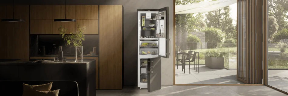 Das Bild zeigt eine geöffnete Kühl-Gefrierkombination zwischen einer Küchenzeile aus Holz und einem zu einer Terrasse hin geöffneten großen Fenster. Das stilvolle Ambiente ist in dunklen Tönen gehalten. 