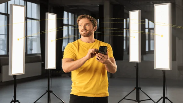 Ein Mann in gelbem T-Shirt und schwarzer Hose steht in einem Raum mit großen Fenstern. Um ihn herum sind fünf längliche, aufrecht stehende Leuchten auf Stativen angeordnet. Er hält ein Smartphone in den Händen und blickt leicht zur Seite. Zwischen den Leuchten sind gelbe Linien eingezeichnet, die eine Verbindung darstellen.